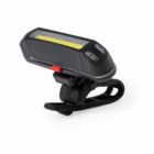 A black bicycle light with an LED strip, mounted on a clip. The light has a rectangular shape with a transparent front and a red button on the side. Its designed for attachment to a bike for visibility and safety.