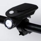 A black bicycle headlight mounted on a handlebar. The light is turned on, emitting a bright beam. The headlight has a sleek design with a prominent button on top.