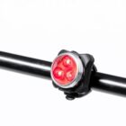 A small, round red bike light with three LEDs is mounted on a black handlebar against a plain white background.