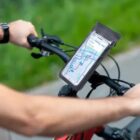 Person riding a bike with a smartphone mounted on the handlebars. The phone displays a map with a route, likely for navigation. The cyclist is on a paved path with grass visible in the background.