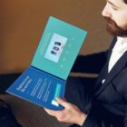 A person in a suit holding an open booklet with a screen displaying images and text. The left page is titled Experience things like never before, with more text and icons, set on a table or desk.