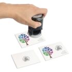 A hand stamps a certificate of authenticity on an art print. Three prints are visible, featuring a colorful tree logo with text reading Art Prints, Matt Stewart, and 1st of Print.