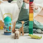 A child in green overalls builds a tall tower with colorful stacking blocks. Wooden animal toys, including a giraffe, elephant, and crocodile, are nearby. A blue and green water bottle with playful designs sits in the foreground.