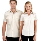 A man and a woman wearing matching beige short-sleeve button-up shirts with black pants stand side by side against a plain white background. Both shirts feature front pockets with flaps.