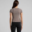 A person with long brown hair is standing with their back to the camera, wearing a short-sleeved, grayish-brown shirt and dark jeans. The background is plain and light-colored.