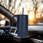 A large navy travel mug with a straw and handle sits in a car cup holder. The background shows a sunset through thin, bare trees.