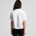 A person with long, dark hair is standing with their back to the camera, wearing a short-sleeve white shirt and black pants. The background is plain and gray.