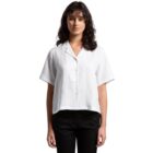 A person with long, dark hair stands facing the camera, wearing a short-sleeved, button-up white shirt and black pants, set against a plain white background.