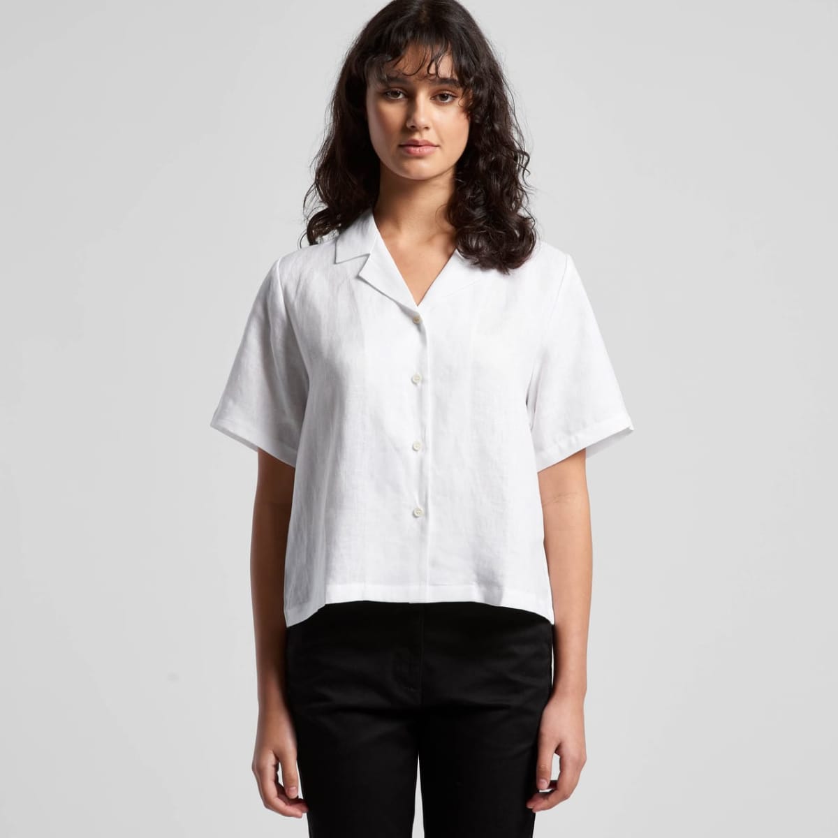 A person stands against a plain gray background, wearing a white short-sleeved button-up shirt and black trousers. The individual has long, dark hair and a neutral expression.