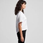 A person with shoulder-length dark hair is standing in profile against a gray background. They are wearing a short-sleeved white shirt and black pants.