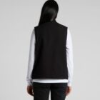 A person with long dark hair is facing away from the camera, wearing a black vest over a white long-sleeve shirt and black pants, standing against a plain light gray background.