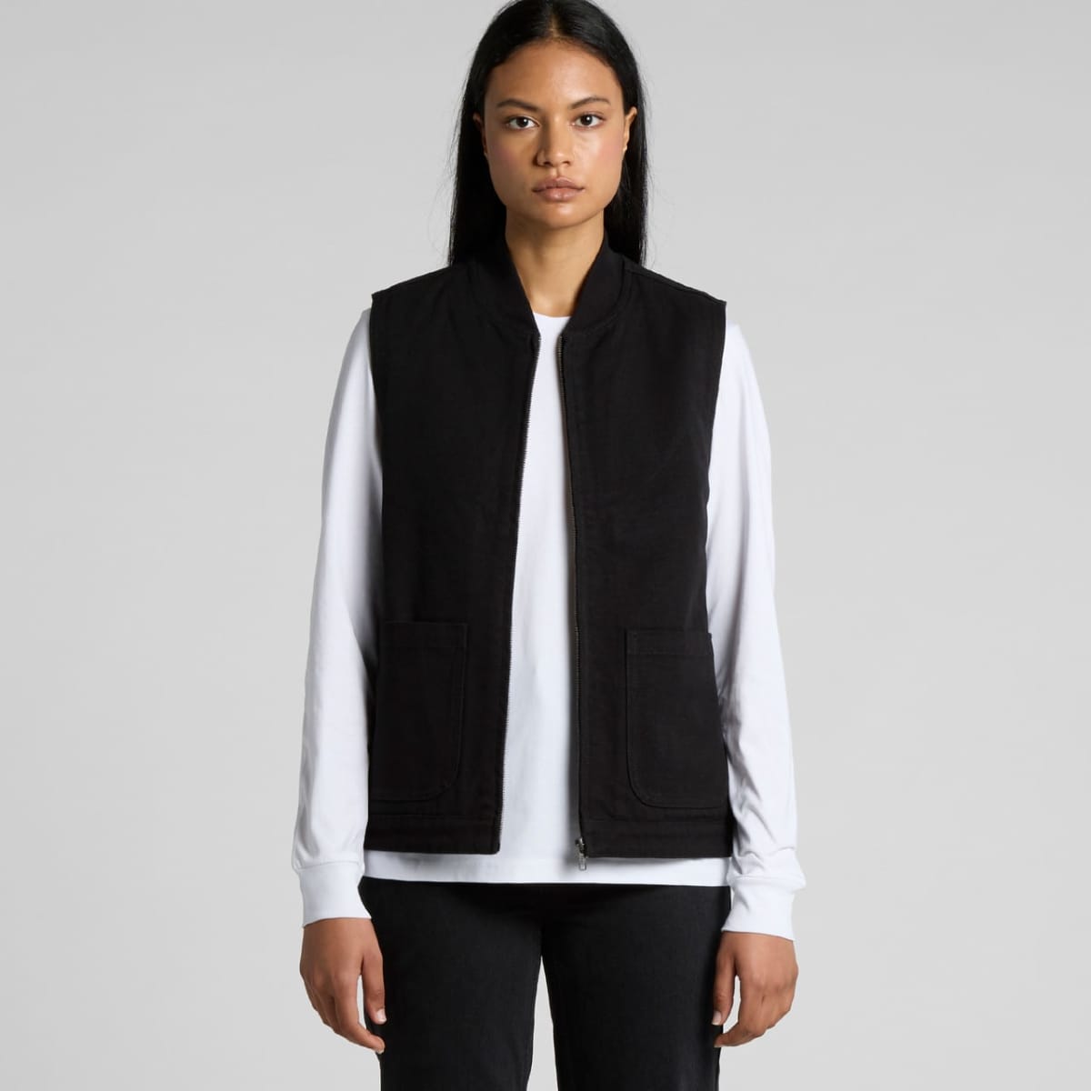 A person with long dark hair wears a black vest over a white long-sleeve shirt and black pants. They stand against a plain gray background, facing the camera directly.
