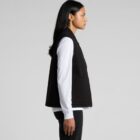Person with long black hair stands in profile against a gray background, wearing a black vest over a white long-sleeve shirt and black pants.