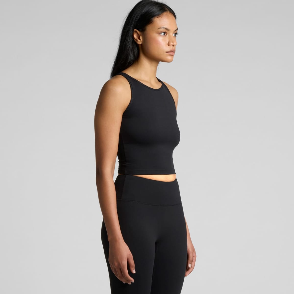 A person wearing a black sleeveless top and black leggings is standing sideways against a plain gray background. They have long dark hair and are looking forward.