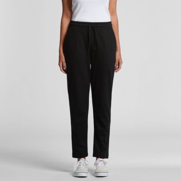 A person wearing a white sleeveless top and black jogger pants stands against a plain gray background. They are also wearing white sneakers.