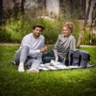 A man and a woman sit on a blanket in a grassy area, having a picnic. They are smiling and wearing casual clothes. A cooler and food items are visible on the blanket. Trees and greenery surround them, creating a peaceful outdoor setting.