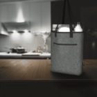 A gray fabric tote bag with black handles stands on a wooden counter in a dimly lit kitchen. The bag contains a rolled-up newspaper, a white plate, and a silver insulated bottle. The background features a modern kitchen with lights.