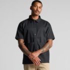 A man wearing a short-sleeved, black button-up shirt and beige pants stands against a plain gray background. He has tattoos on both arms and is looking slightly to the side with his hands clasped in front.
