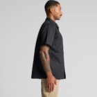 A person with short hair, wearing a black short-sleeve collared shirt and beige pants, stands in profile against a plain, light-gray background. Their arm is decorated with colorful tattoos.