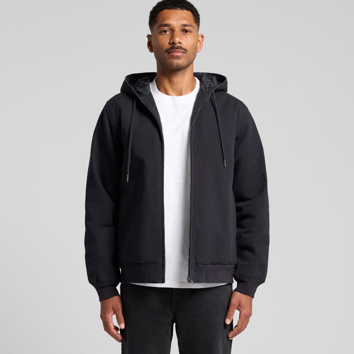 A person wearing a black zip-up hoodie over a white T-shirt and black pants stands against a plain gray background. The hoodie is worn with its hood down.
