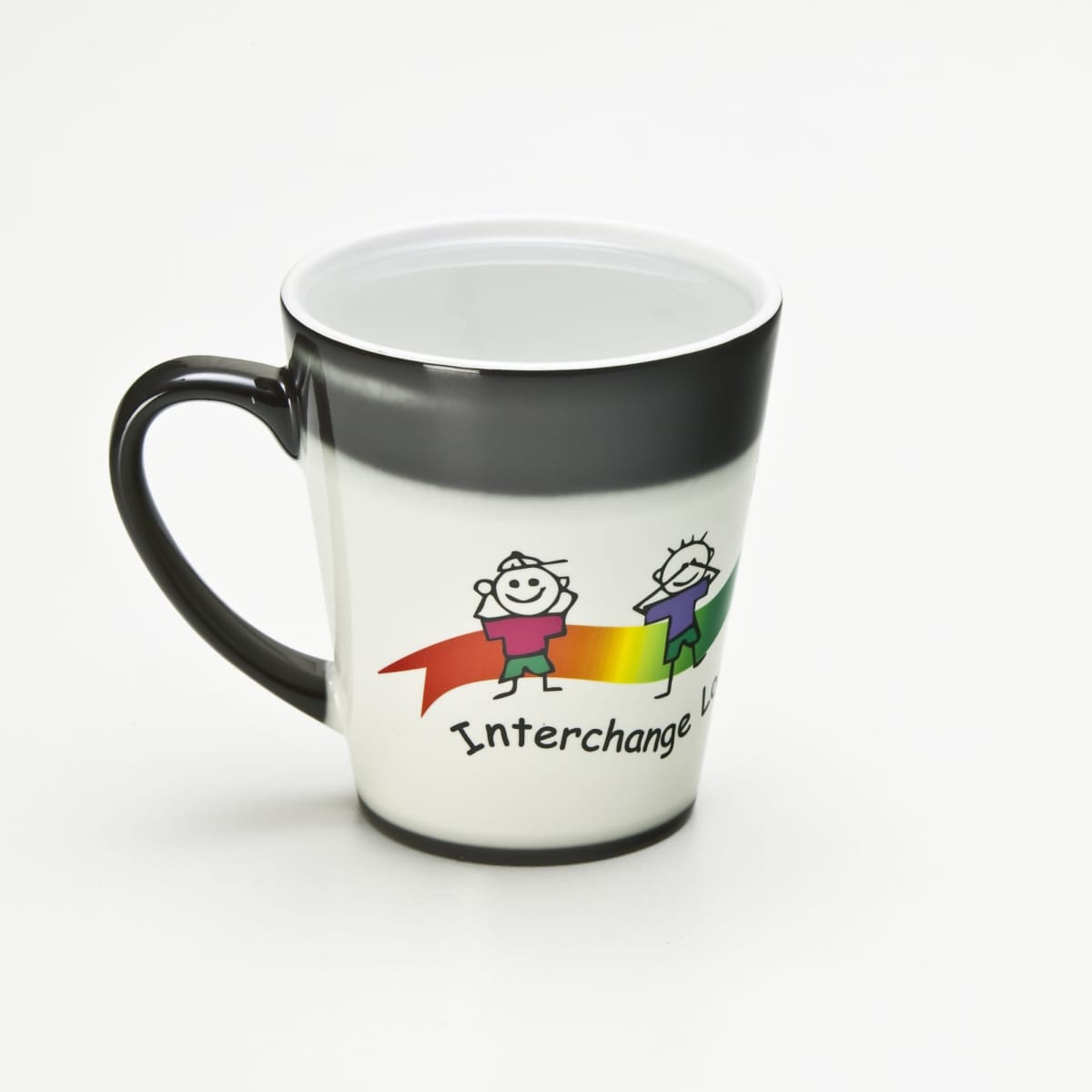 The Latte Magic Mug is a white mug with a black handle and rim, featuring two smiling stick figures holding a colorful ribbon. Interchange U.K. is printed below, revealing surprises with each latte.