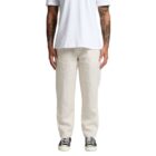 A person wearing a white T-shirt and beige pants stands facing the camera. Their arms are visible and feature tattoos. They are also wearing black and white sneakers. The background is plain white.