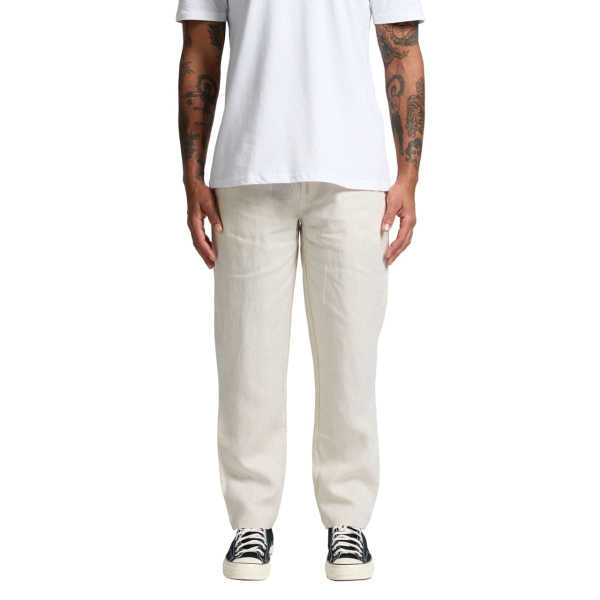 A person wearing a white T-shirt and beige pants stands facing the camera. Their arms are visible and feature tattoos. They are also wearing black and white sneakers. The background is plain white.