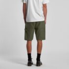 A person is standing with their back to the camera, wearing a white T-shirt, green cargo shorts, black socks, and brown shoes. The background is a plain, light gray.
