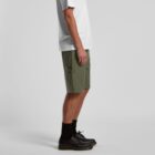 A person stands sideways in a minimalistic setting, wearing a white T-shirt, green shorts, black socks, and black shoes. The background is plain light gray.