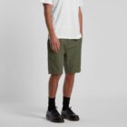 A person wearing a white T-shirt, olive green shorts, black socks, and black shoes stands against a plain gray background. The persons hands are in their pockets.