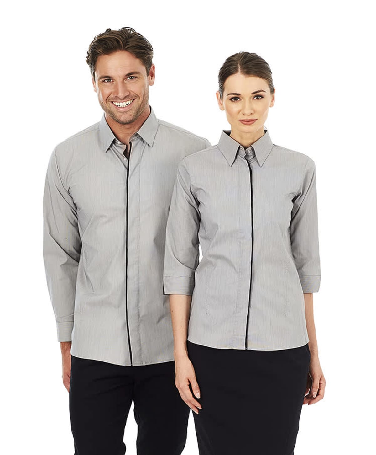 A man and a woman stand side by side, both wearing light gray button-up shirts with black pants. The man has his hands in his pockets, and the woman stands with her arms at her sides. They are smiling and facing forward.