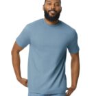 A man with a beard is smiling and wearing a light blue T-shirt and jeans. He has his hands on his hips and stands against a plain white background.