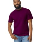 A man with a beard is smiling, wearing a burgundy T-shirt and light blue jeans. He is standing against a plain white background, with one hand in his pocket.