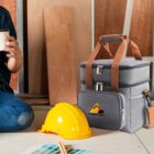 A person, partially visible, sits on a floor holding a drink next to a gray lunch bag with brown straps and a yellow hard hat. Wooden panels are in the background, suggesting a construction or workshop setting.