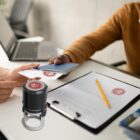A person hands a document to another across a desk. A stamp and a clipboard with a pen are visible on the table. A laptop is open in the background. The document has a red seal.