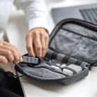 A person is organizing a gray tech pouch on a desk. The pouch contains cables and a gadget with USB ports. A laptop is partially visible in the background. The person is wearing a white shirt.