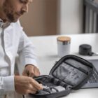 A man in a white shirt sits at a desk, organizing items inside a black zippered case. A silver travel mug and a laptop are on the desk. The scene conveys a sense of organization and preparation.