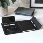 A sleek, organized desk setup featuring a leather portfolio with a notepad, pen, phone holder with a smartphone, and card slots. A potted plant and a closed portfolio are in the background, creating a modern and tidy workspace.
