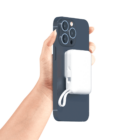 A hand holding a smartphone with a white, compact accessory attached to the back. The accessory has a short cable. The phone has a dark colored case and a triple-lens camera.