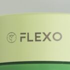 Close-up of a light green product with the word FLEXO and a logo featuring a circular design with a line and dot elements. Below is a darker green base. The background is plain.