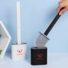 A person holds a black silicone toilet brush above its matching black holder. Next to it, theres a white toilet brush with a white holder. Both holders have the logo LoveHouse printed on them.