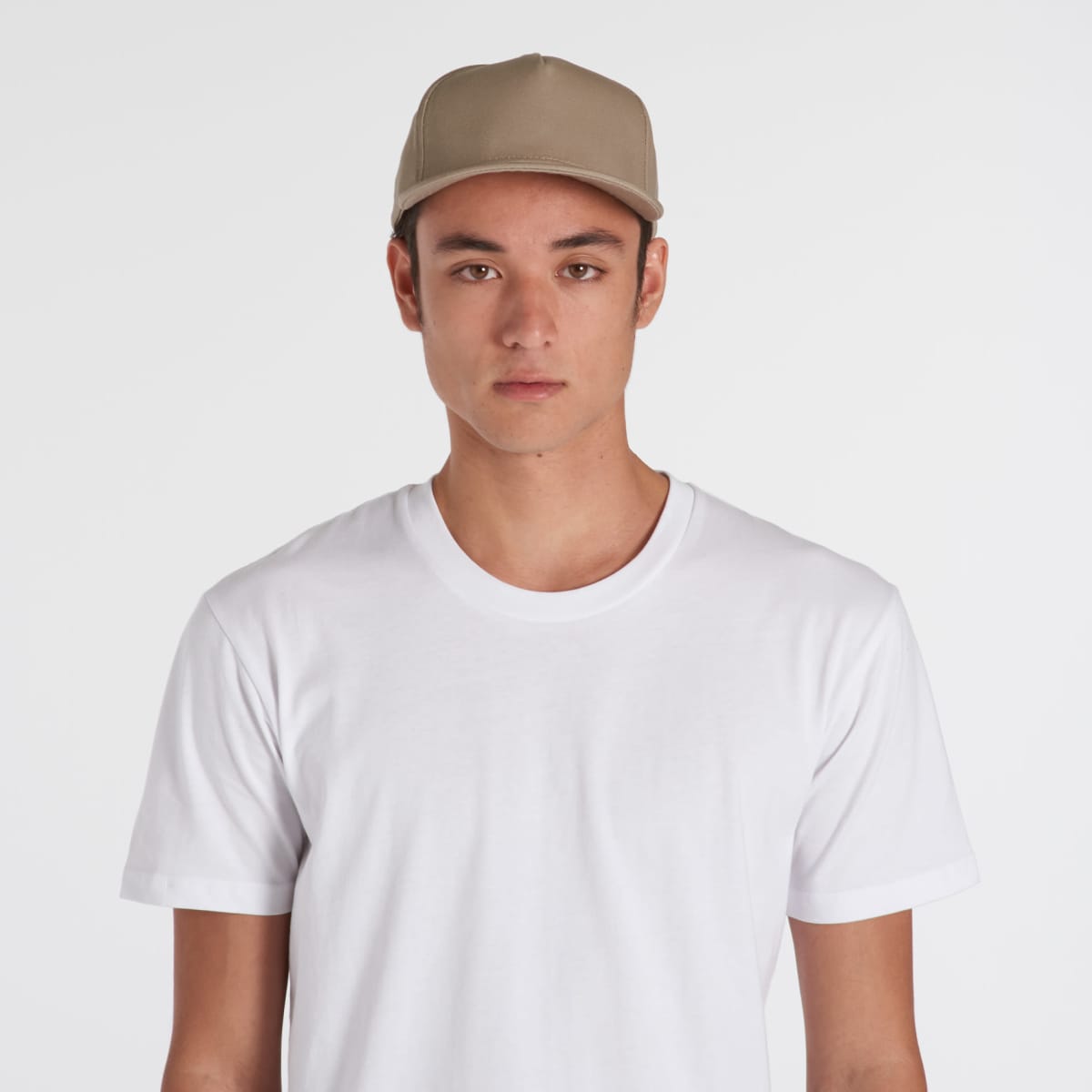 A person wearing a Tommy Cap in beige and a plain white T-shirt stands against a plain white background, looking directly at the camera.