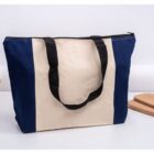 A canvas tote bag with a zippered top. It features a beige center panel and navy blue side panels. The bag has black handles and is placed on a white surface, with wooden boards in the background.