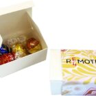 A white gift box with assorted Lindor chocolates in blue, red, gold, and black wrappers by Lindt is open. Next to it is a closed REMOTE box with a yellow and pink abstract design. Both products are part of the 4-Pack Lindt Ball Choc Box.