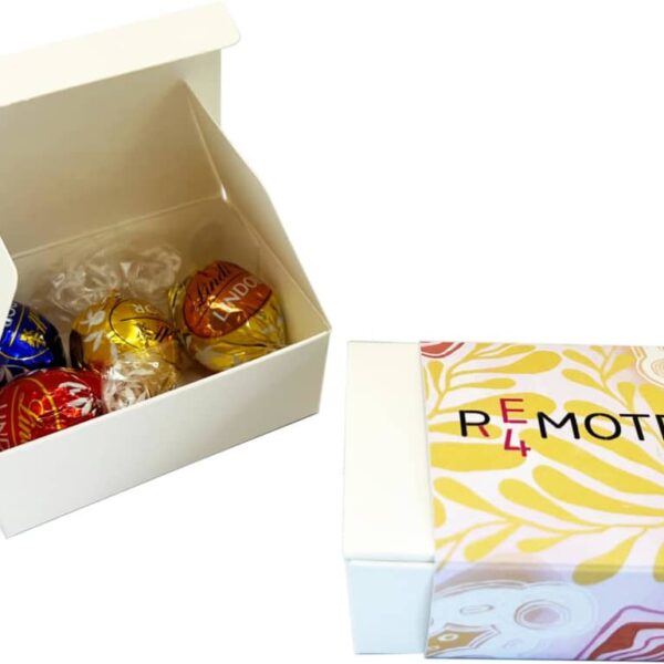 A white gift box with assorted Lindor chocolates in blue, red, gold, and black wrappers by Lindt is open. Next to it is a closed REMOTE box with a yellow and pink abstract design. Both products are part of the 4-Pack Lindt Ball Choc Box.