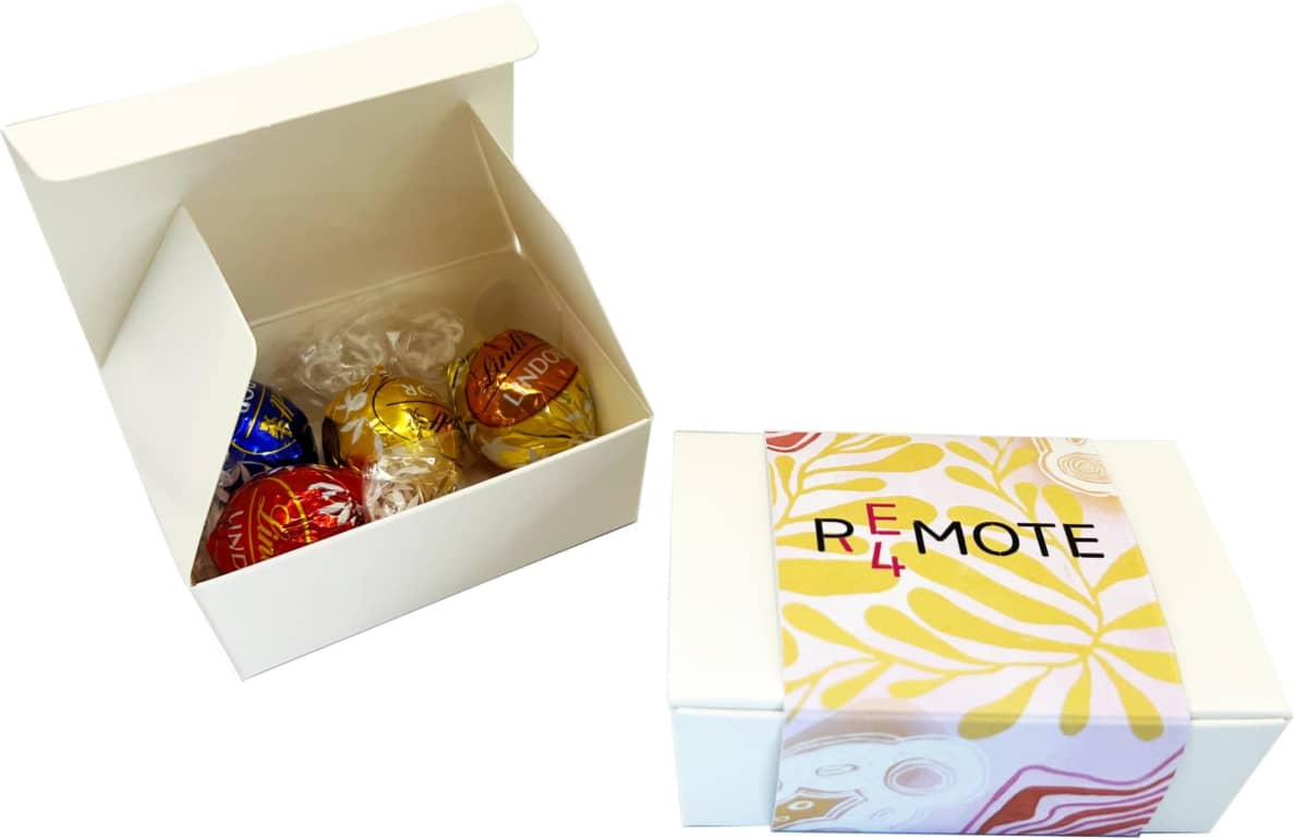 A white gift box with assorted Lindor chocolates in blue, red, gold, and black wrappers by Lindt is open. Next to it is a closed REMOTE box with a yellow and pink abstract design. Both products are part of the 4-Pack Lindt Ball Choc Box.