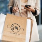 A person in a beige coat holds a smartphone and several shopping bags. One bag is brown with SM Susannah Mae printed on it in black. The background is blurred, emphasizing a focus on shopping and style.