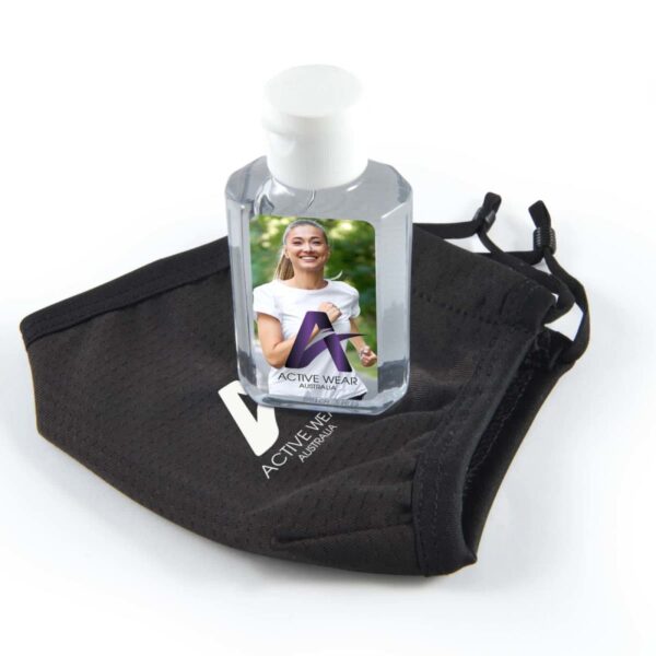 A small bottle of hand sanitizer with a label featuring a smiling woman stands on a black face mask. Both items have an Active Wear Australia logo. The mask has black ear loops, and the sanitizer bottle has a white cap.