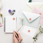 A persons hand holds a pen above a spiral notebook labeled Aero. Surrounding the notebook are envelopes, a business card, flowers, and small gift containers, all also featuring the Aero logo. The setting is bright and clean.