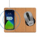 A wireless charging mat holds a smartphone displaying a charging icon alongside a time and date. A wireless mouse sits next to the phone on the mat. A charging cable is connected to the mat. The mat has a cork-like texture.